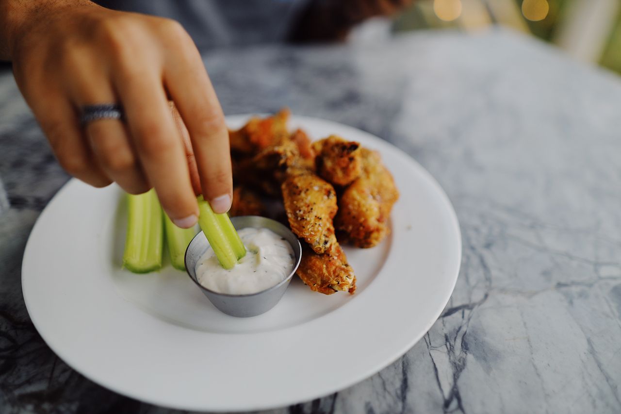Close-up of a hand dipping celery stick in ranch sauce next to crispy chicken wings.